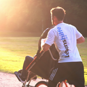 Ein Mann in weißem T-Shirt mit Lebenshilfe-Logo schiebt einen Menschen im Rollstuhl